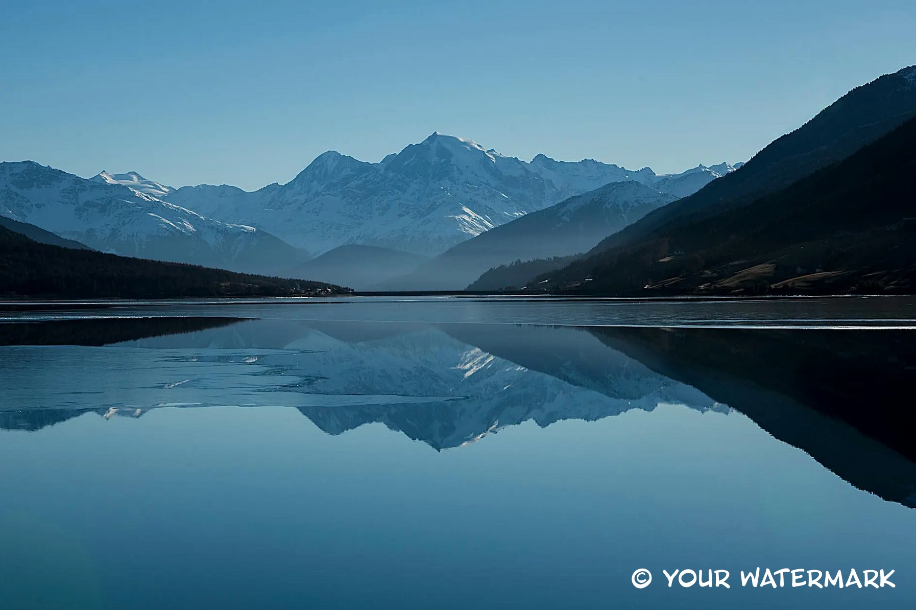 Snow-capped mountains reflected in a lake with a small text watermark in the lower corner